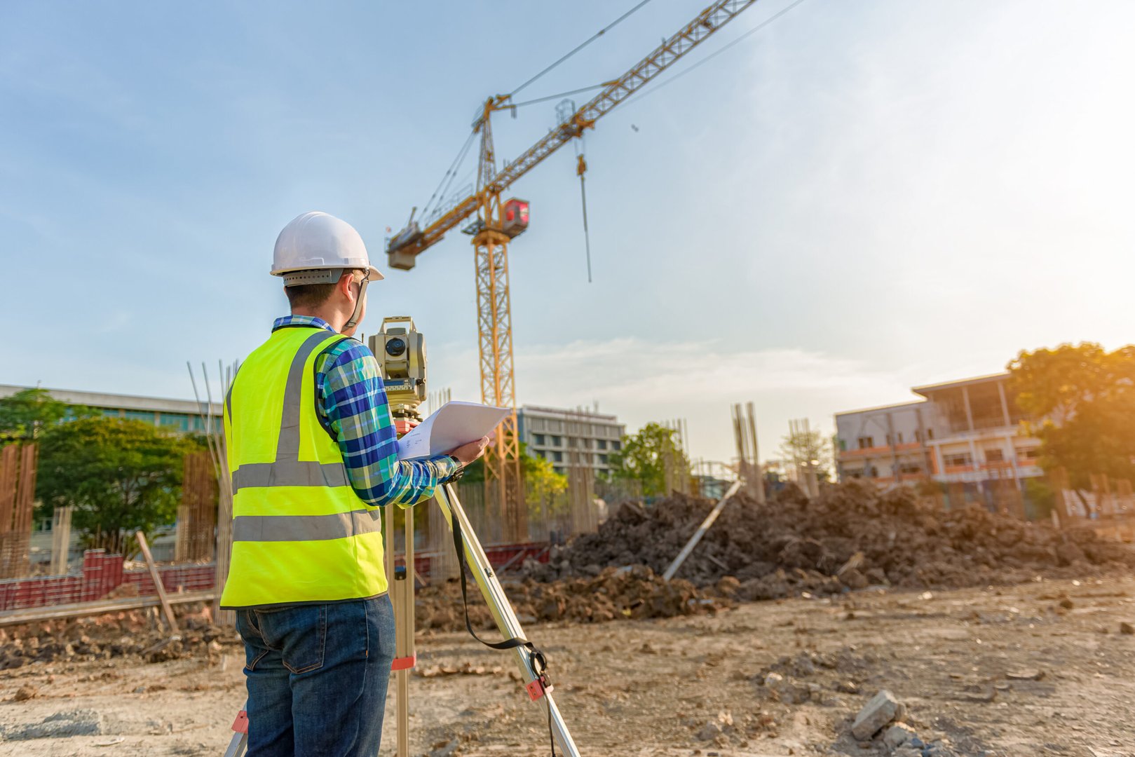 Man in high-visibility jacket surveying a project with a theodolite with a crane in the background